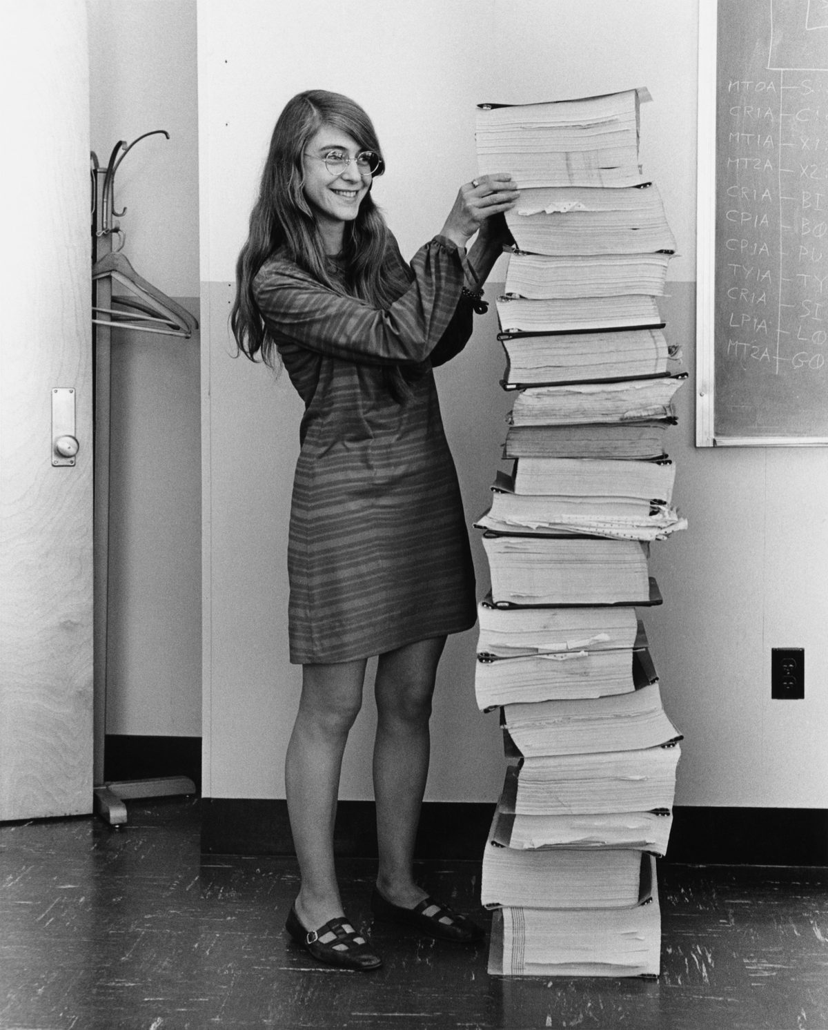 Margaret Hamilton, souriante, pose la main sur une pile de listings de programme aussi haute qu'elle